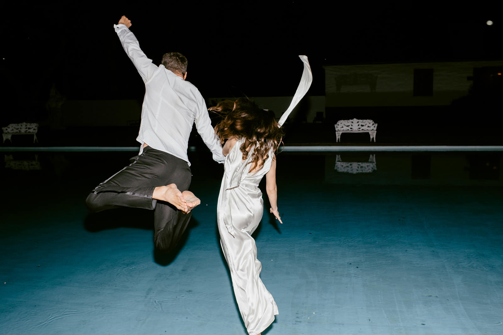 Nighttime wedding photo of a bride and groom jumping into the pool in their wedding attire at Häringe Slott. Captured by wedding photographer Karin Lundin. Taken with direct flash for a dynamic effect.