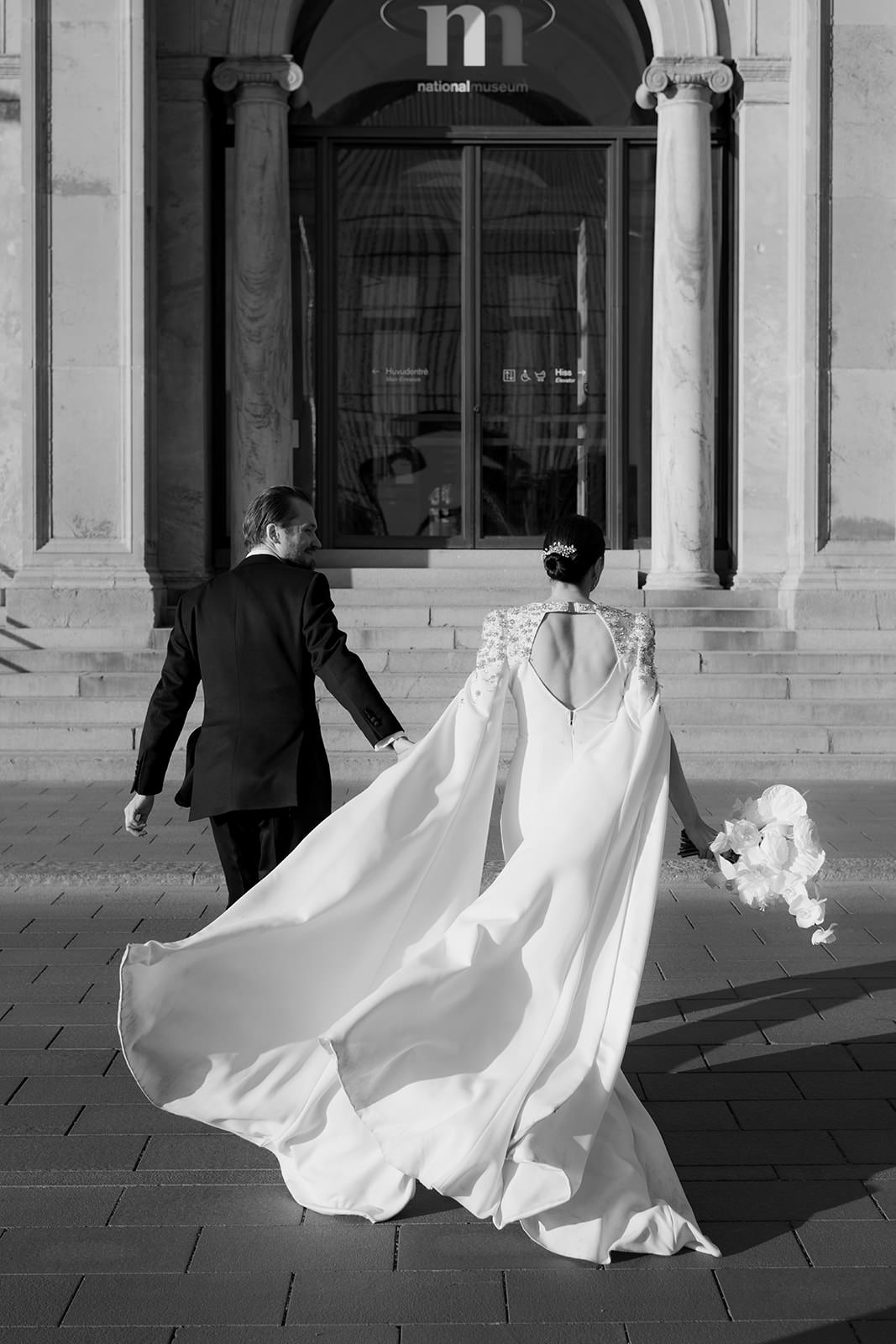 Black and white photo of a bride and groom holding hands as they walk toward Nationalmuseum in Stockholm for their wedding dinner. The bride wears a dress with a cape flowing in the wind. Captured by wedding photographer Karin Lundin. Planned by Events by Sara.