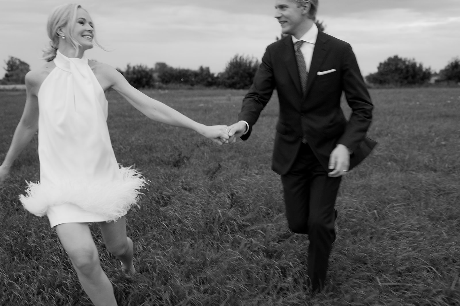 Black and white photo of a bride and groom running across a field at dusk. The bride wears a short white dress with fringe and feather details at the hem. Captured in Torekov, Skåne, by wedding photographer Karin Lundin.