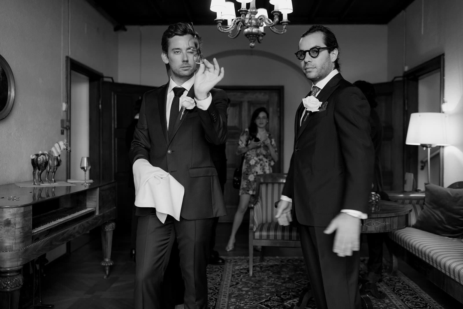 Black and white photo of the groom and best man in black suits at Högberga Gård. Planned by WeddingPlanner Stockholm. One polishes his glasses while the other looks into the camera wearing sunglasses. Captured by wedding photographer Karin Lundin during the preparation before the wedding ceremony.