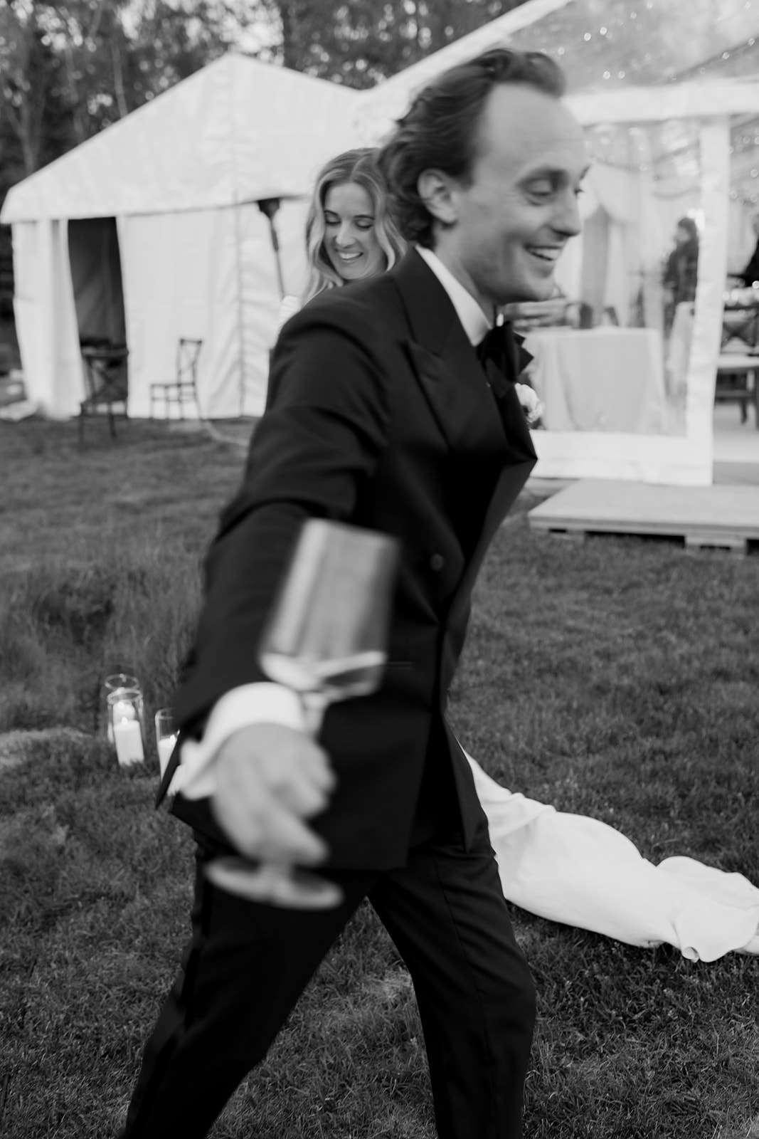 Black and white photo of a bride and groom holding hands and spinning together on a summer evening in the Stockholm archipelago. Captured by wedding photographer Karin Lundin. The groom, closest to the camera, holds a glass of champagne. Captured with a slow shutter speed for motion blur. Planned by WeddingPlanner Stockholm.