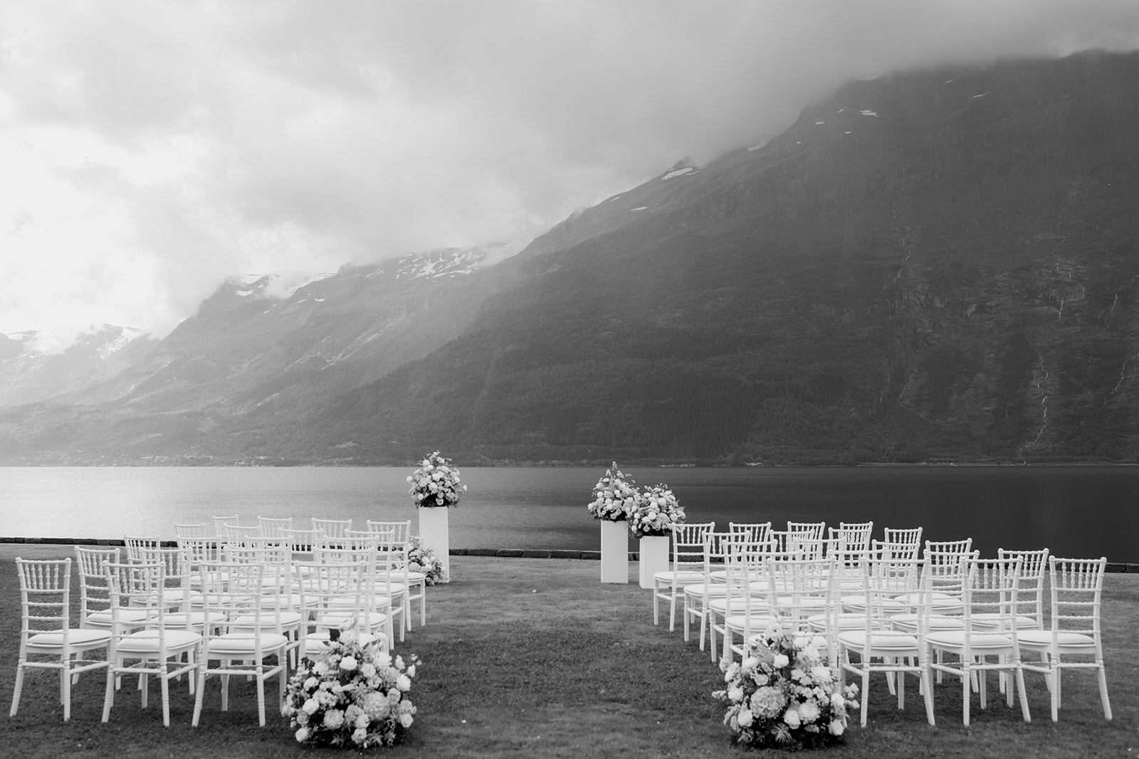 Black and white photo of a wedding ceremony setup by the Norwegian fjord at Ullensvang Hotel. Captured by wedding photographer Karin Lundin. White chairs and floral arrangements by Bloomster i Bergen are displayed on pedestals. Planned by Weddingsbyemma.