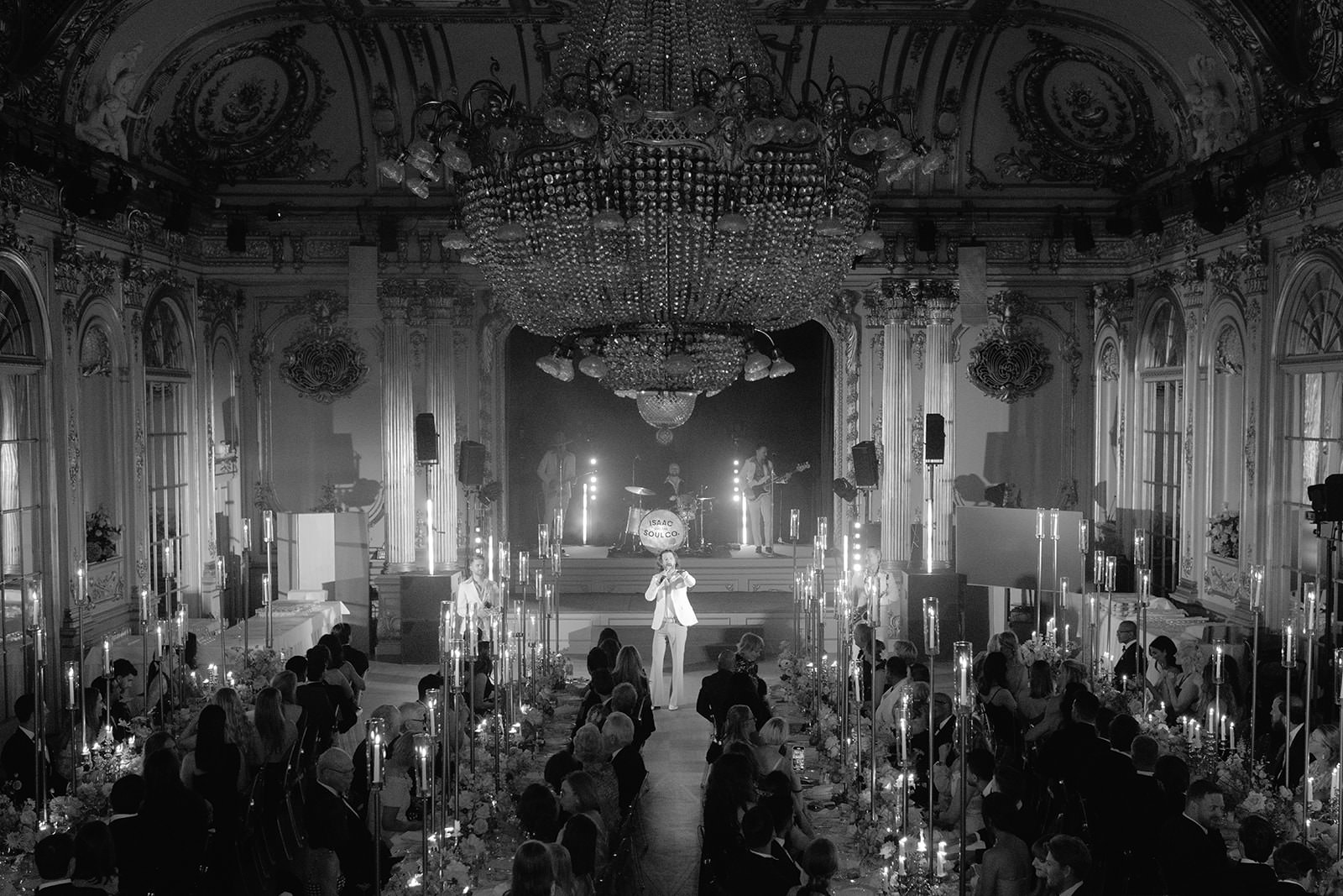 Black and white photo of the Hall of Mirrors at Grand Hôtel in Stockholm, captured from the balcony. Captured by wedding photographer Karin Lundin. The band Isac and the Soul Company performs as guests stand and applaud at their tables. Planned by WeddingPlanner Stockholm.