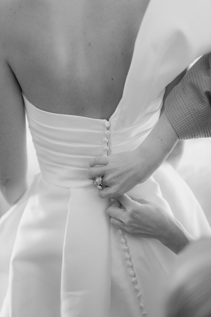 Wedding at Norrvikens trädgårdar 47 Black and white close-up of the wedding dress buttons being fastened, ring on the hand