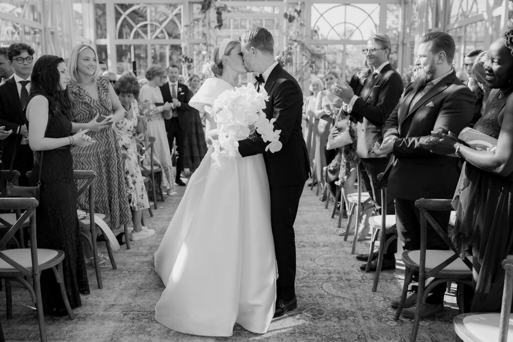 Wedding at Norrvikens trädgårdar 48 Black and white: the couple kisses in the aisle of Victoriahuset as guests stand and applaud