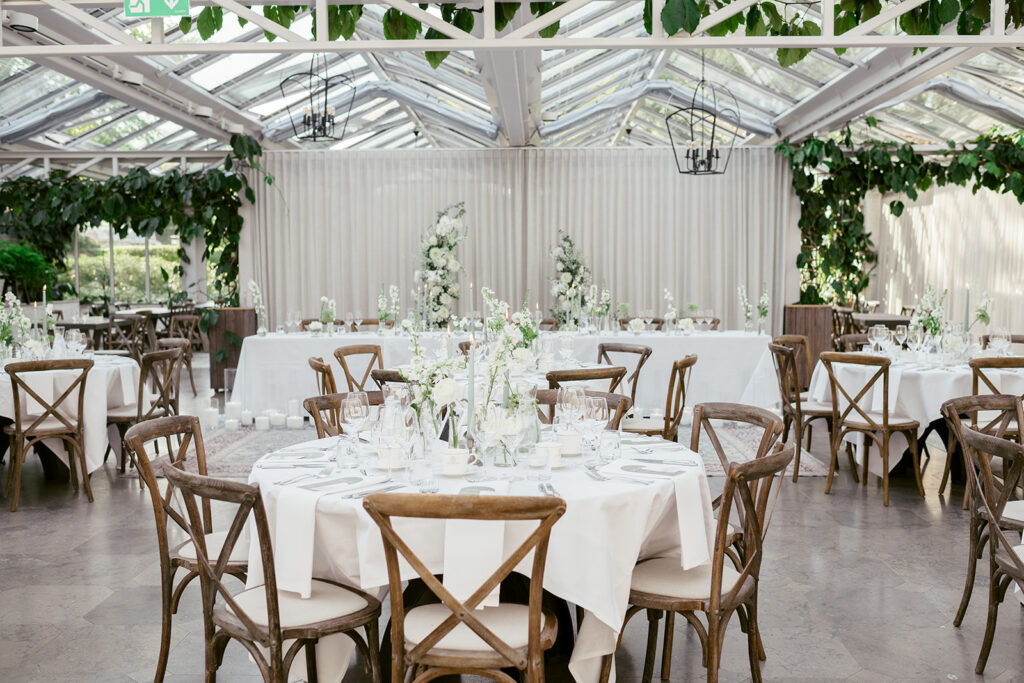 Wedding at Norrvikens trädgårdar 57 Dinner tables with white cloths and wooden chairs under a glass roof with hanging greenery