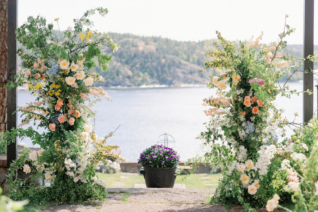 Slottsbröllop i Sverige: slott och herrgårdar 12 Floral ceremony arch with pink and peach roses framing a view of the Stockholm archipelago at Högberga Gård