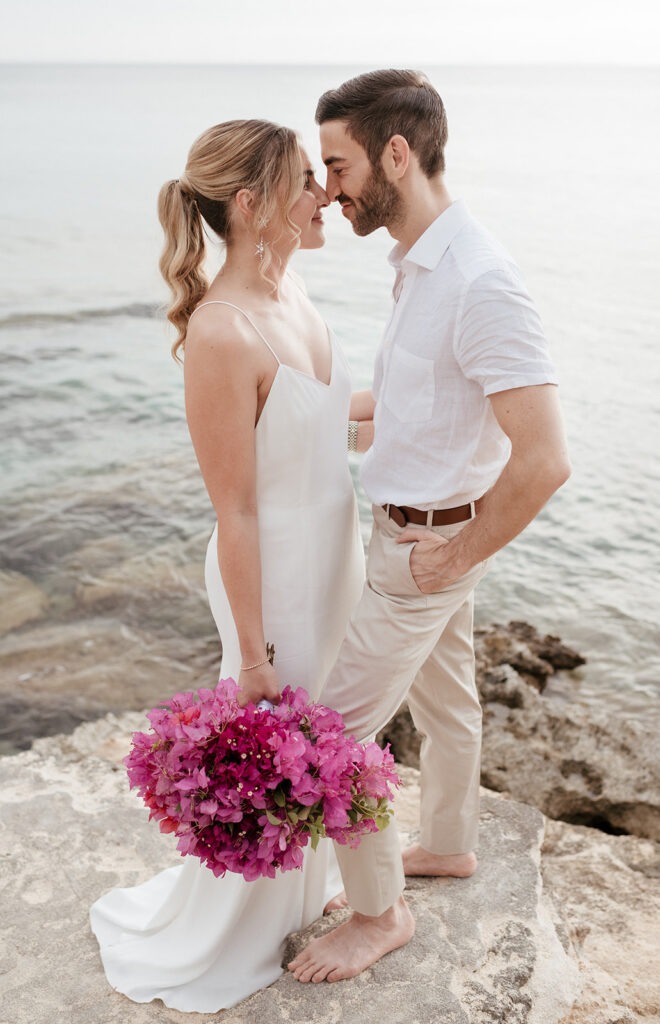 Bride and groom forehead to forehead by the sea on Kefalonia, magenta bougainvillea bouquet in her hand