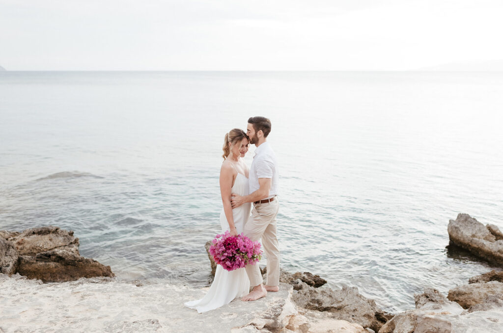 Wedding couple standing on the rocks at the shore of Kefalonia, pale sea behind them