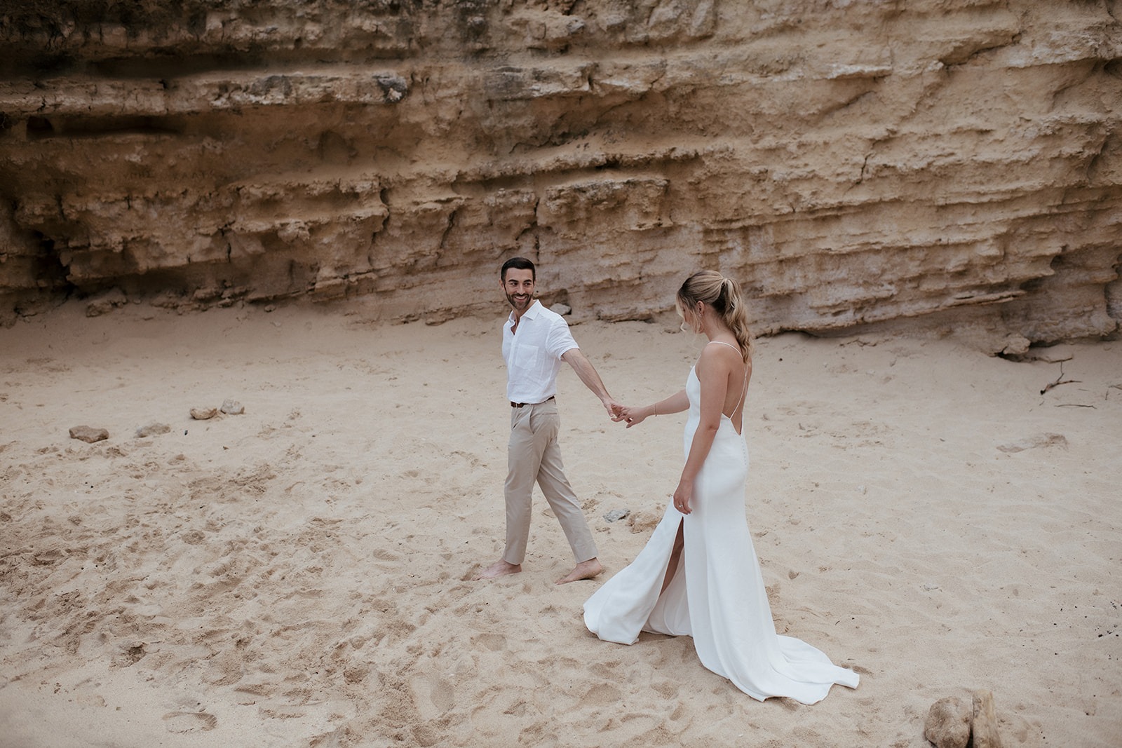 Bride and groom walking hand in hand on a small beach under a sandstone cliff, Kefalonia