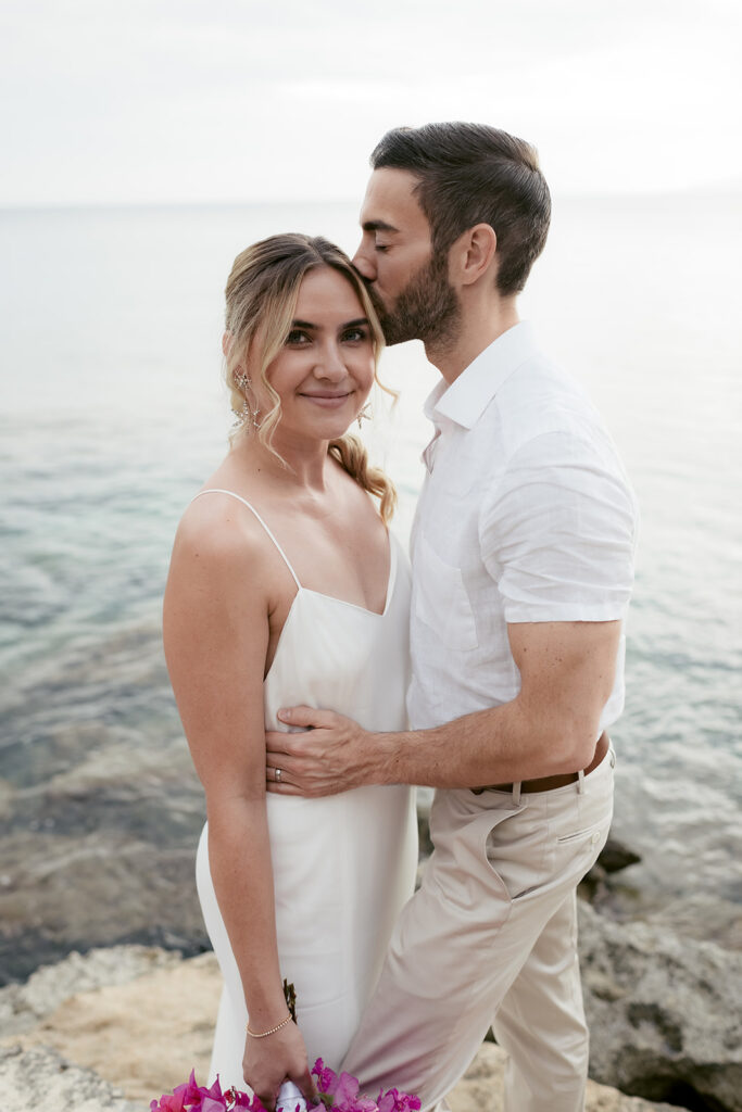 Bride smiling at the camera while the groom kisses her hair, bougainvillea bouquet, Kefalonia