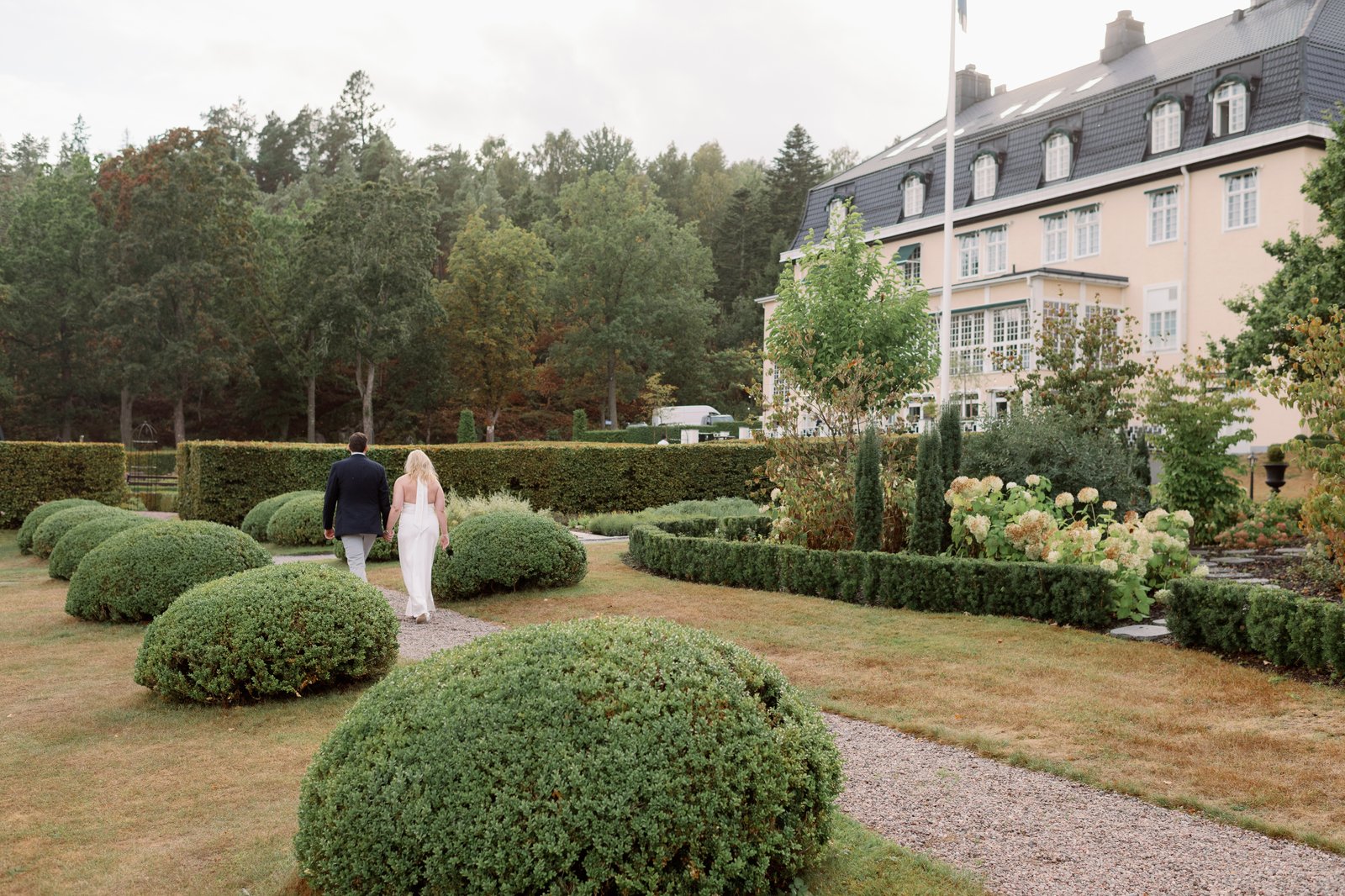 Bröllop på Villa Fridhem: en fotografs guide 1 Brudparet står i den formella trädgården bland klippta buxbomsklot, Villa Fridhem i bakgrunden