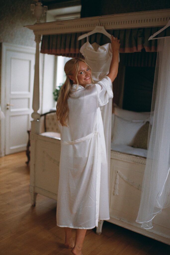 Wedding at Thorskogs Slott 37 The bride in a robe hangs up her dress by a four-poster bed, shot on 35mm film