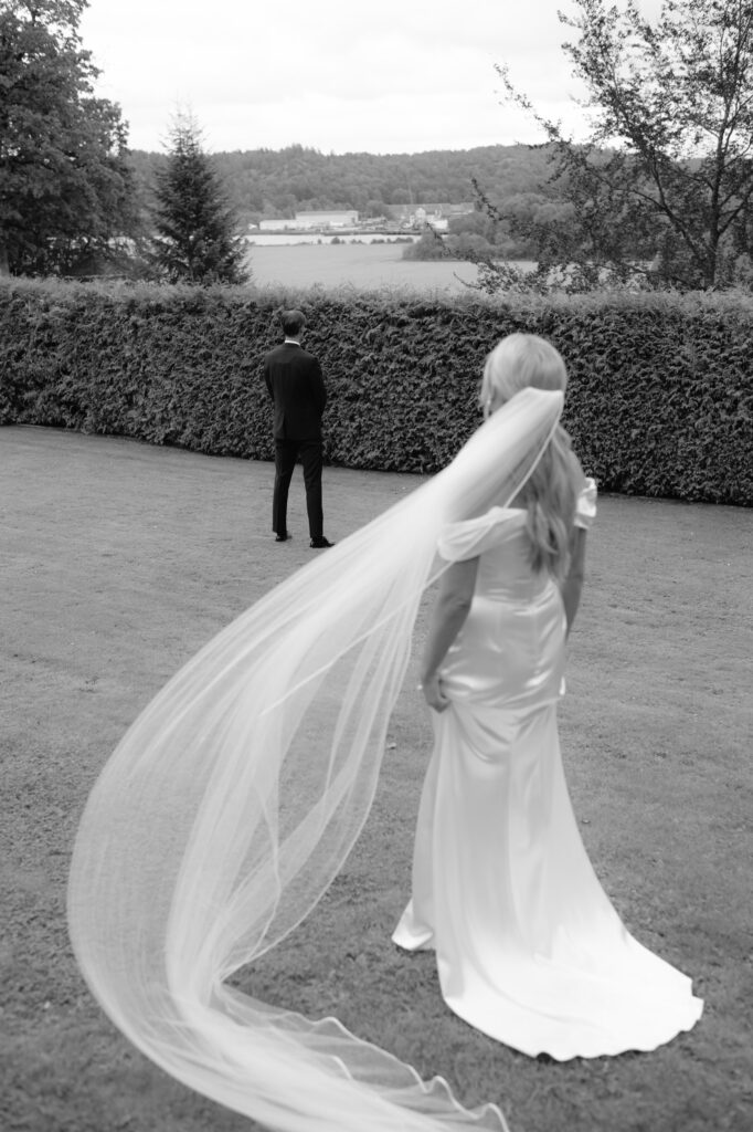 Wedding at Thorskogs Slott 29 Black and white: the bride walks across the lawn towards the groom who stands with his back turned, her veil lifting in the wind