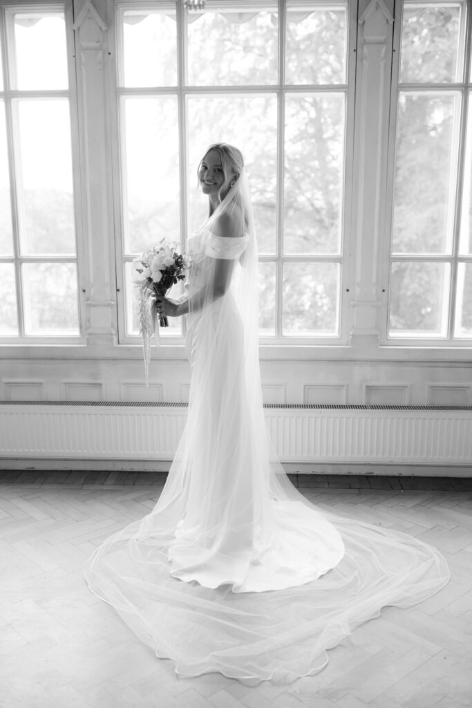 Wedding at Thorskogs Slott 27 Black and white: the bride stands by a tall window with her bouquet, the train spread across the floor