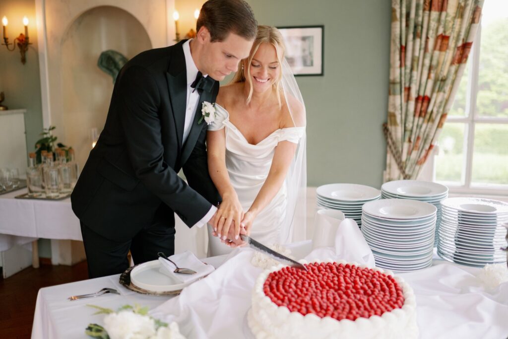 Wedding at Thorskogs Slott 25 The couple cuts the wedding cake topped with red berries, stacked plates beside it