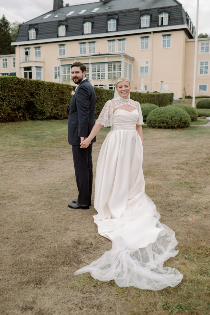 The couple holds hands in front of Villa Fridhem, the veil spread across the grass