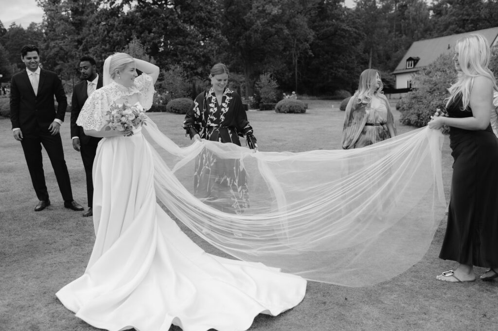 Black and white: guests hold up the long veil of the bride on the lawn