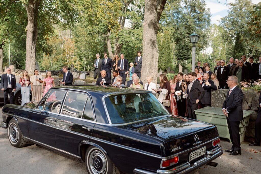 Guests gather around a black vintage car under the trees