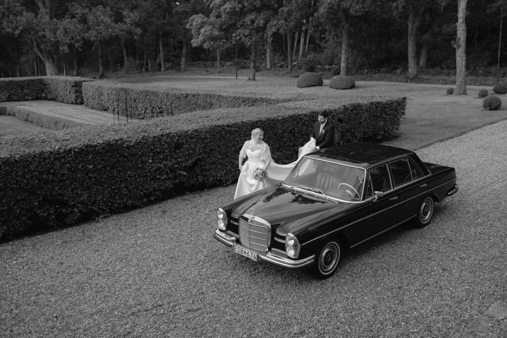 Bröllop på Villa Fridhem: en fotografs guide 5 Svartvitt: brudparet i den svarta veteranbilen på grusvägen genom trädgården