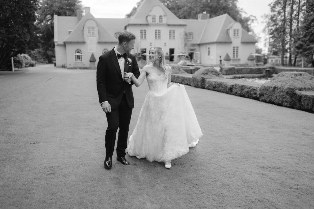Wedding at Norrvikens trädgårdar 42 Black and white: the couple walks along the gravel drive in front of a large white house, the bride lifting her dress