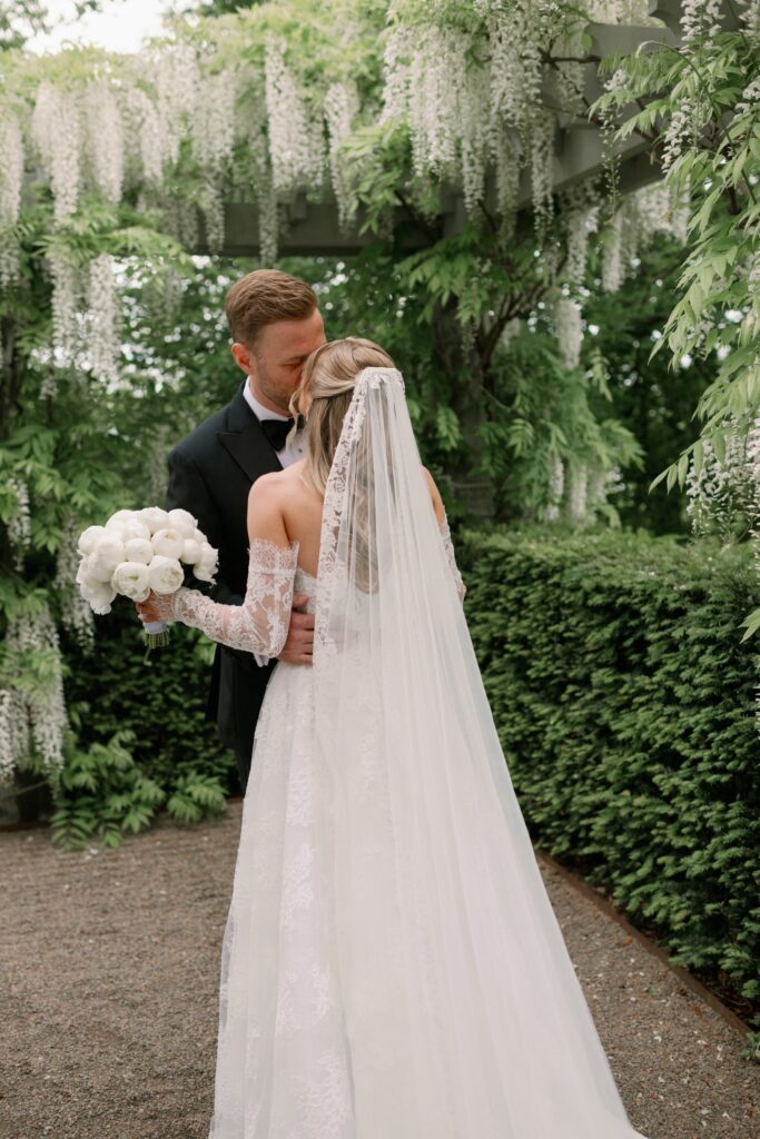 Wedding at Norrvikens trädgårdar 41 The groom kisses the bride on the cheek under the wisteria, a white bouquet in her hands