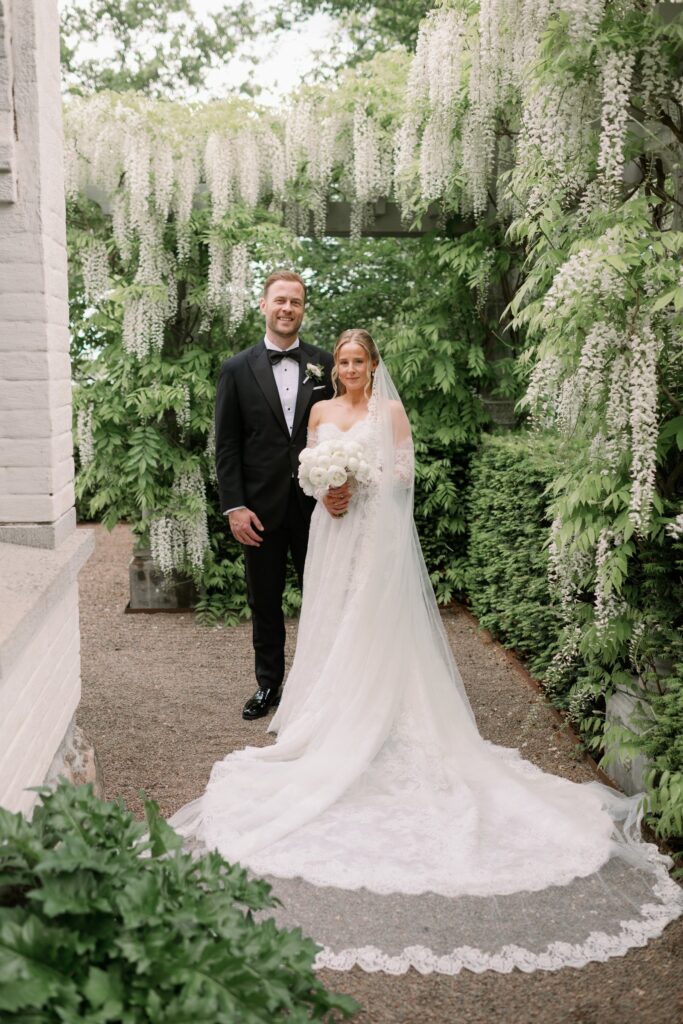 Wedding at Norrvikens trädgårdar 39 The couple stands under white wisteria smiling at the camera, the bride holding a white bouquet