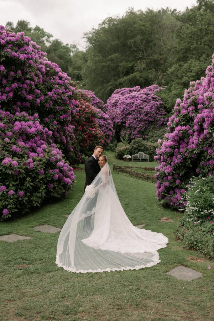 Wedding at Norrvikens trädgårdar 20 The couple stands on the lawn between purple rhododendrons, the train spread across the grass