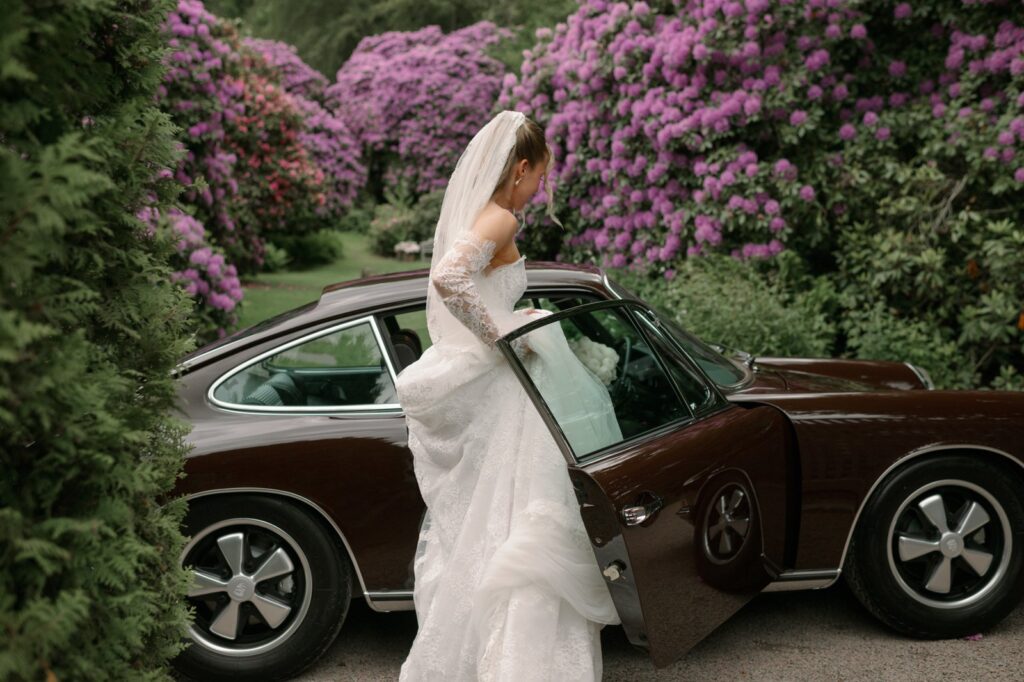 Wedding at Norrvikens trädgårdar 6 The bride steps out of a dark brown vintage car in front of purple rhododendrons