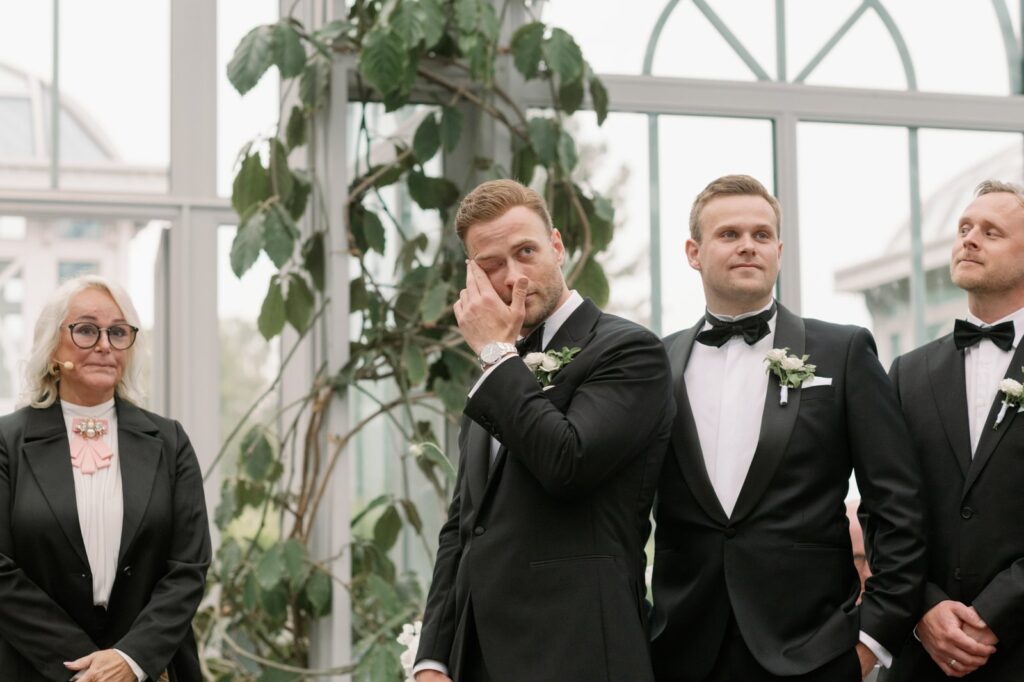 Wedding at Norrvikens trädgårdar 31 The groom wipes away a tear during the ceremony, the officiant and friends in tuxedos beside him
