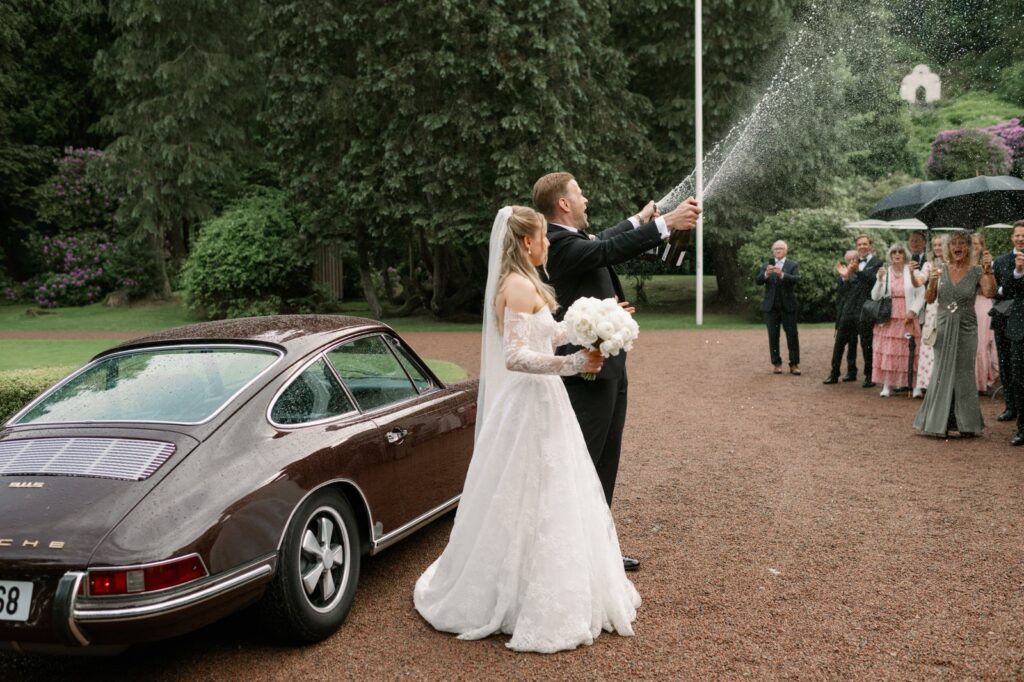 Wedding at Norrvikens trädgårdar 26 The groom sprays champagne beside the bride by the vintage car, guests with umbrellas behind them