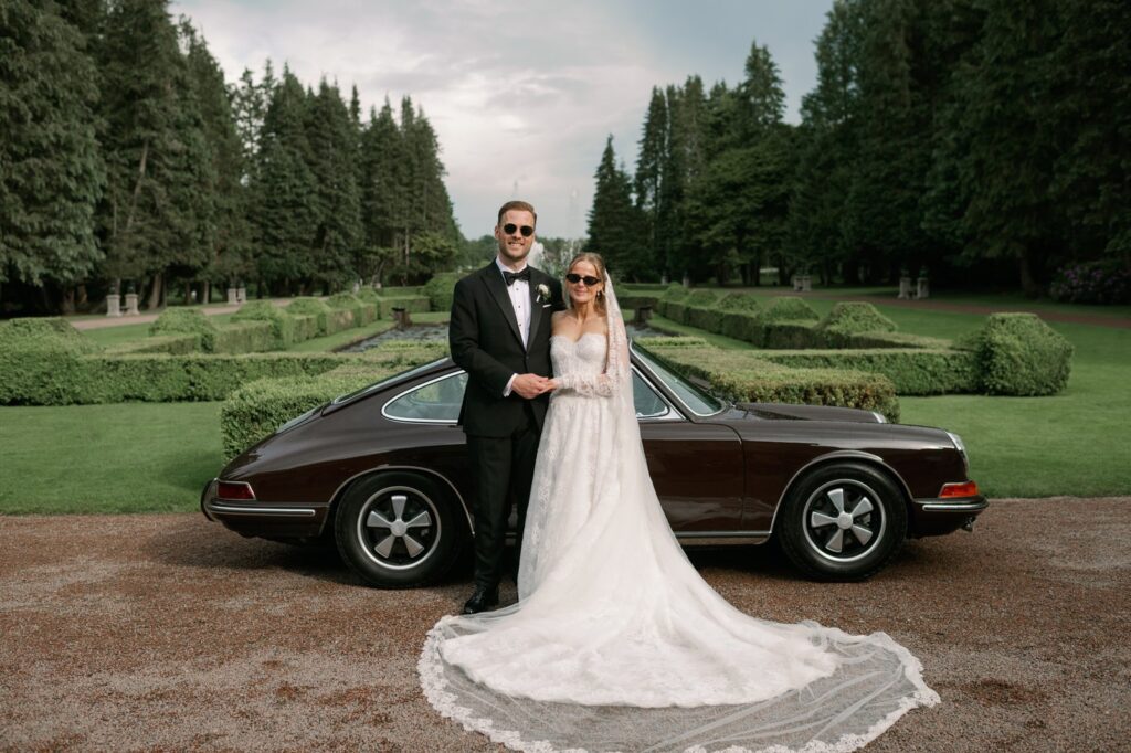 Wedding at Norrvikens trädgårdar 24 The couple in sunglasses in front of a dark brown vintage car by the formal garden with clipped hedges