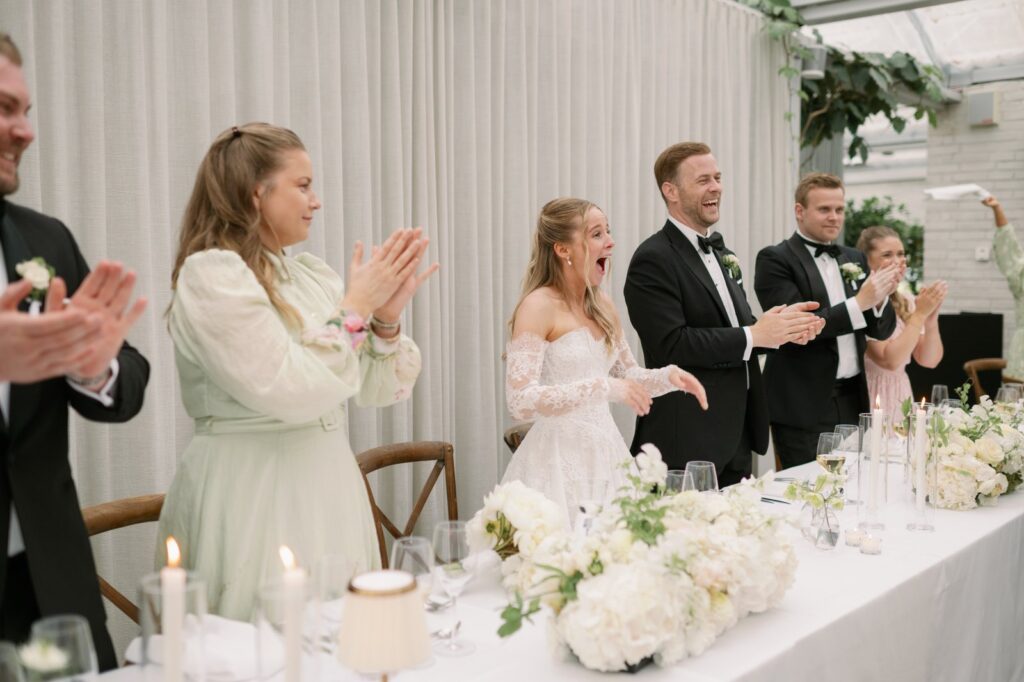 Wedding at Norrvikens trädgårdar 17 The bride and guests laughing and applauding at the long table with white flowers and candles