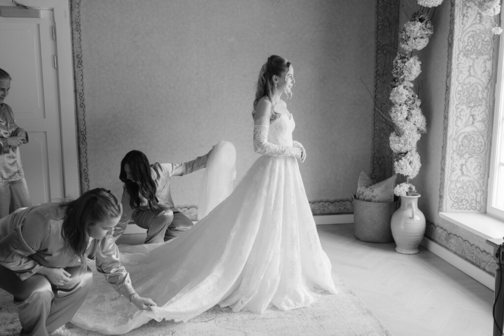 Wedding at Norrvikens trädgårdar 8 Black and white: two women adjust the train of the dress in a bright room while the bride gets ready
